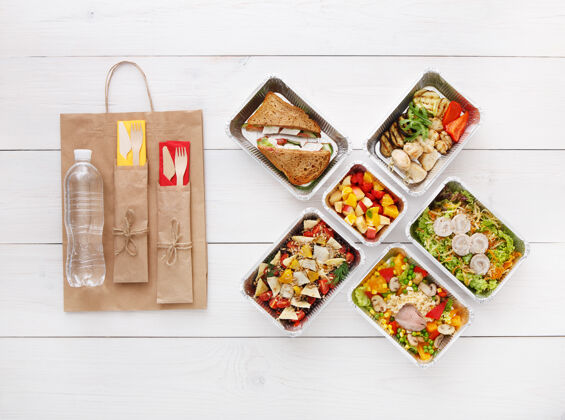 烹饪健康食品配送外卖食品铝箔盒中的蔬菜 肉类和水果 餐具 水和牛皮纸包装俯视图 平放在白色木头上 有复印空间包装纸外卖