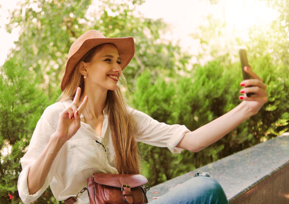 年轻休闲风格的金发女人戴着毡帽在户外自拍坐在城市公园里公园电话自拍