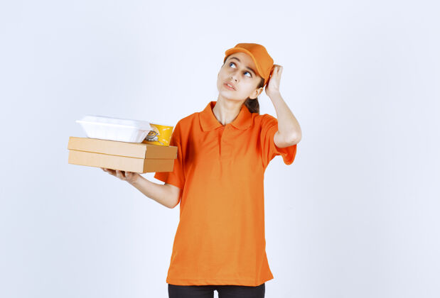员工身着橙色制服的女快递员手里拿着一个纸板箱 一个塑料外卖箱和一个黄色的面杯 神情迷茫 若有所思姿势购物犹豫