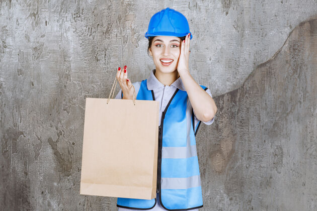 促销身穿蓝色制服 头戴安全帽的女工程师拿着购物袋 看上去很惊讶快乐服装货物