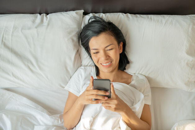 阅读微笑迷人的亚洲女性在白色床上使用智能手机放松年轻房子