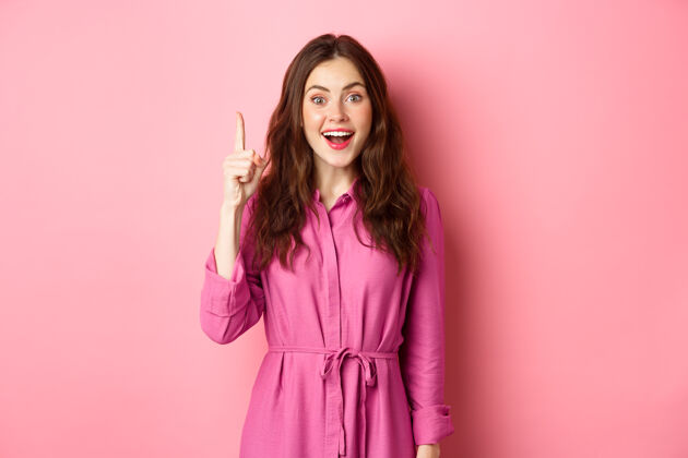 请兴奋美丽的女孩化妆 有一个想法 举起手指在尤里卡标志和微笑 说她的计划 站在粉红色的墙壁上黑发积极女性
