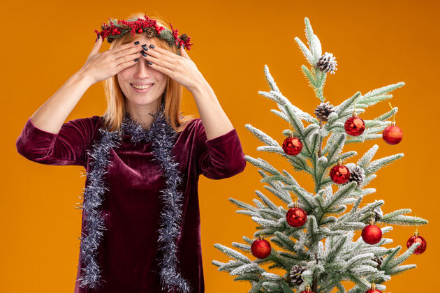 圣诞节站在圣诞树旁微笑的年轻漂亮女孩 身穿红色连衣裙 脖子上戴着花环 眼睛蒙着 手孤立在橙色的墙上手附近花圈