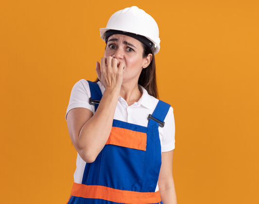 表情一个穿着制服的关心建筑的年轻女人咬着隔离在橙色墙上的钉子人脸感情