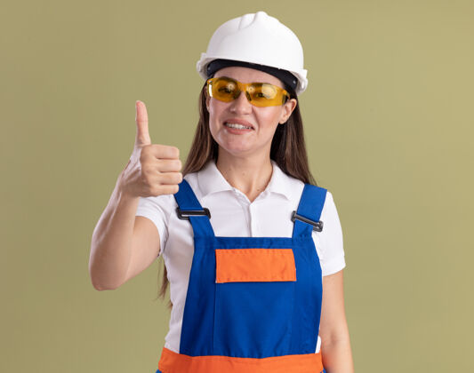 制服身着制服 戴着眼镜 微笑着的年轻建筑工人孤立地站在橄榄绿的墙上 竖起大拇指眼镜衣服站立