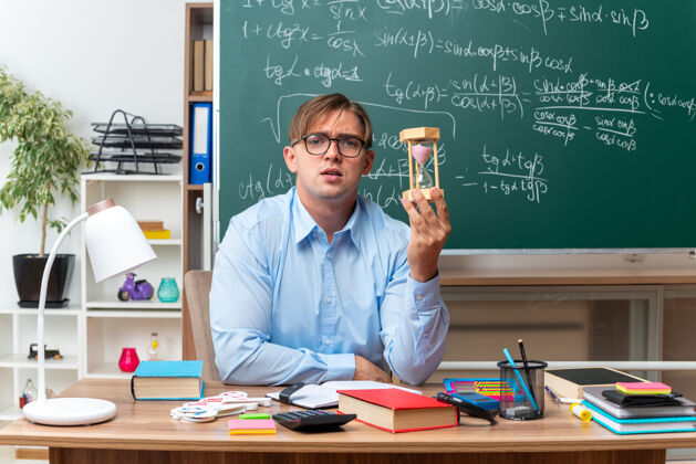 课桌年轻的男老师戴着沙漏眼镜备课 自信地坐在课桌旁 教室的黑板前放着书和笔记教室黑板准备