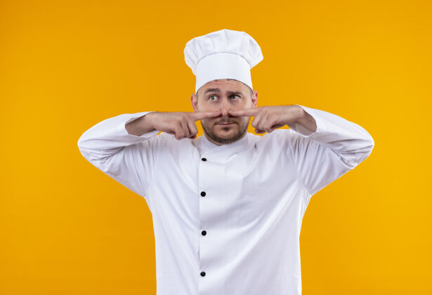 印象印象深刻的年轻帅哥厨师制服把手指放在鼻子上看一边孤立在橙色墙上烹饪帅气手指