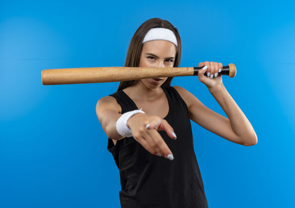 指尖自信的年轻漂亮的运动女孩戴着头带和腕带拿着棒球棒指着蓝色的墙上 看起来孤立腕带年轻抱着