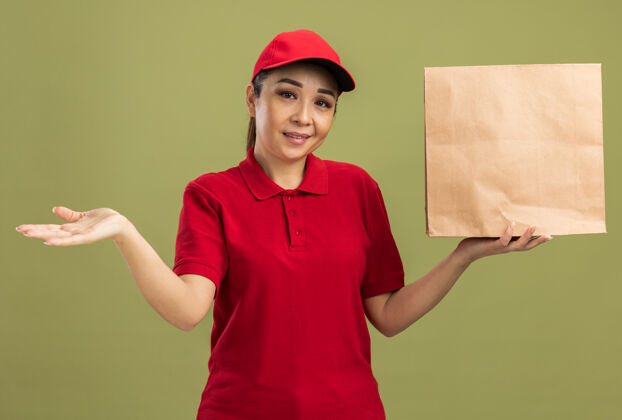 脸快乐的年轻送货员 身穿红色制服 戴着帽子 手里拿着纸包 脸上带着微笑 胳膊站在绿色的墙上手臂举行微笑