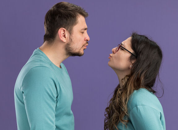 衣服穿蓝色休闲服的年轻漂亮夫妇站在紫色的墙上 要热吻相爱的男人和女人站立男人年轻