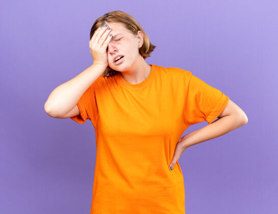 感觉不健康的年轻女子穿着橘色t恤 站在紫色的墙上摸额头感到不适 头晕 感冒额头年轻站立