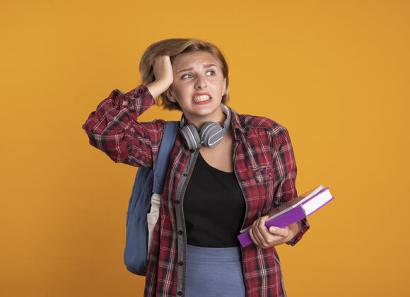 橙色焦急的年轻斯拉夫女学生戴着耳机背着背包把手放在头上拿着书和笔记本穿学生头