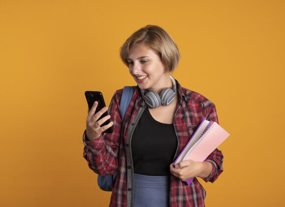 微笑微笑的年轻斯拉夫女学生戴着耳机背着背包拿着书和笔记本看着手机电话空间耳机