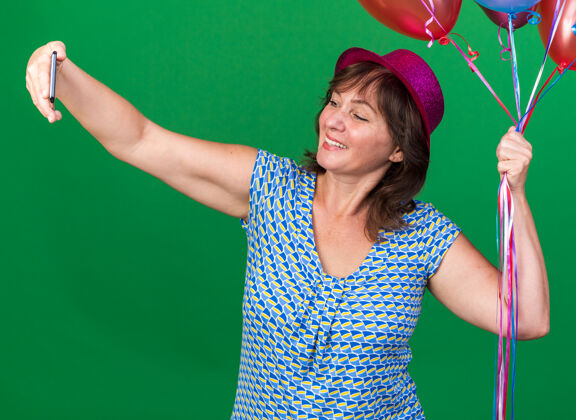 年龄戴着派对帽的中年妇女拿着五颜六色的气球用智能手机自拍快乐欢快的微笑庆祝生日派对站在绿色的墙上中间手机女人