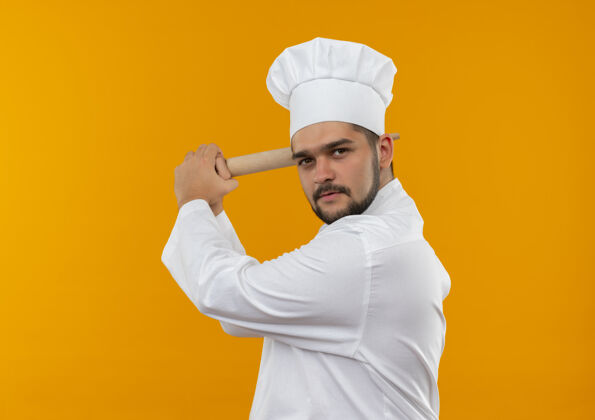 立场自信的年轻男厨师身着厨师制服站在侧视图中拿着擀面杖 准备用复制空间打败孤立的橙色墙壁男性准备年轻