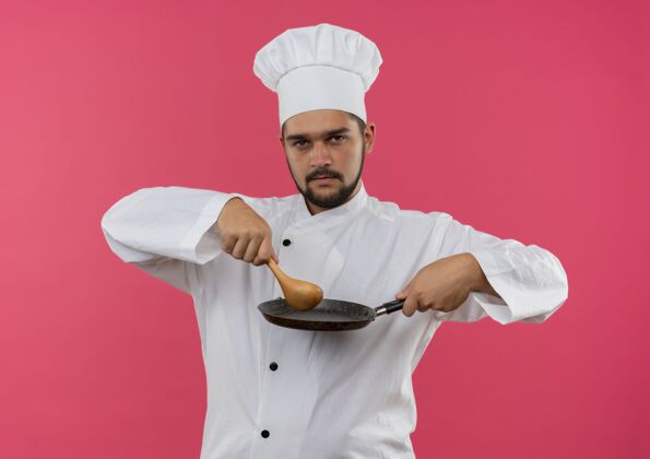平底锅自信的年轻男性厨师身着厨师制服 拿着汤匙和煎锅 隔离在粉红色的墙上制服勺子年轻