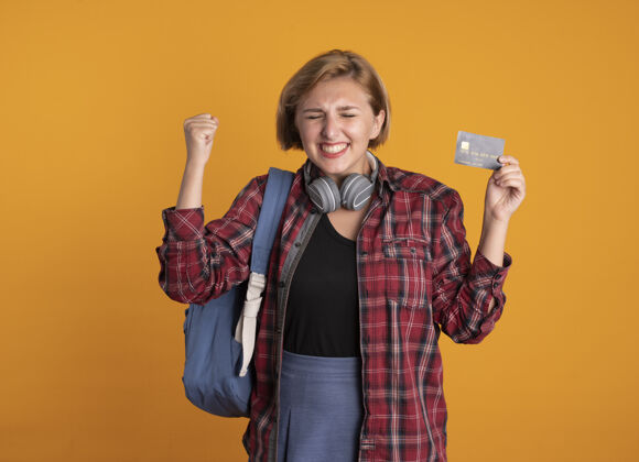 复制兴奋的年轻斯拉夫女学生戴着耳机背着背包站着闭着眼睛举起拳头拿着信用卡背包耳机眼睛