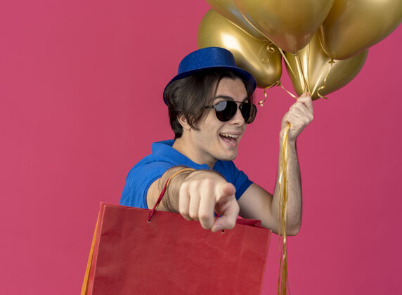快乐一个戴着太阳眼镜 戴着蓝色派对帽 手里拿着氦气球和纸购物袋 指着相机的快乐帅哥指向持有帅哥