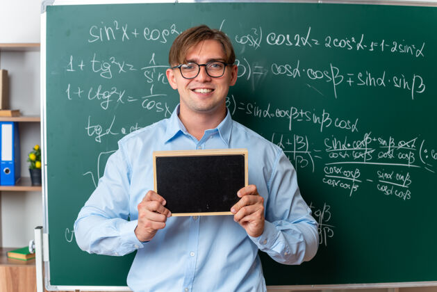 微笑年轻的男老师戴着眼镜 拿着小黑板 面带微笑 自信地站在教室里的数学公式黑板旁年轻数学戴着