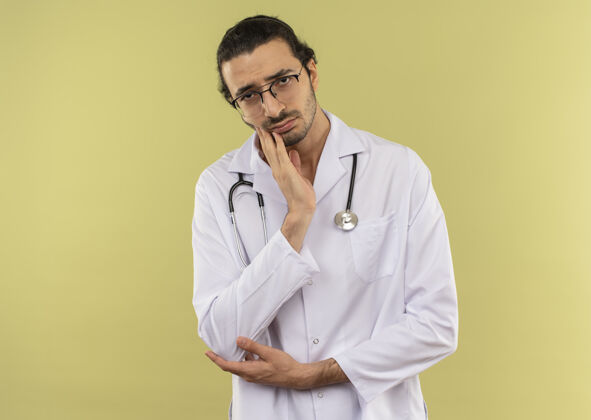 下巴悲伤的年轻男医生 戴着眼镜 穿着白色长袍 听诊器 手放在绿色背景的下巴上听诊器长袍眼镜