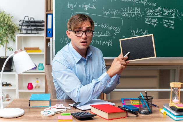 小年轻的男老师戴着眼镜 用小黑板和粉笔讲解课文 自信地坐在课桌旁 教室里黑板前放着书和笔记桌子学校人