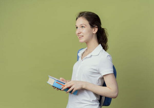垫面带微笑的年轻漂亮的女学生 背着书包站在侧视图里 手里拿着笔记本 笔直地看着书背部绿色橄榄
