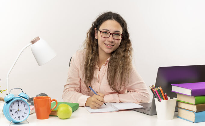 市民面带微笑的年轻漂亮的女学生戴着眼镜坐在课桌旁 拿着学习工具做作业 在白墙上孤立的记事本上写字桌子女生作业