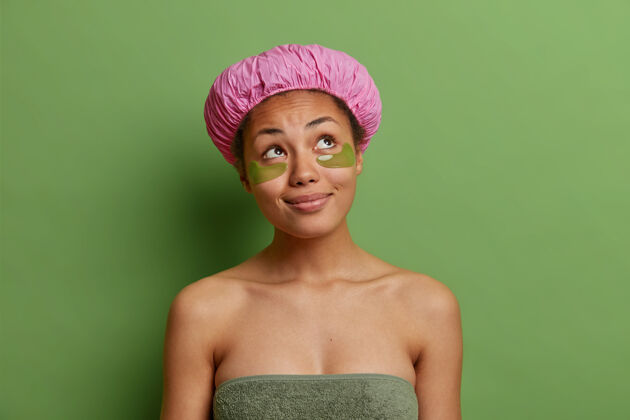 健康体贴的年轻女子裹着浴巾在眼睛下面敷上水凝胶贴片 戴着隔离在绿色墙壁上的浴帽美容年轻清洁