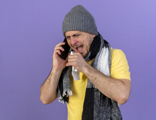 电话恼怒的年轻金发病斯拉夫男子戴冬季帽子和围巾举行电话纸巾谈话恼怒金发纸巾