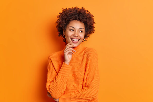 喜悦年轻漂亮的非裔美国女孩一边看一边带着牙齿的微笑注意到令人愉快的事情有着无忧无虑的表情穿着休闲套头衫在生动的橙色墙壁上摆出室内姿势满意毛衣成人