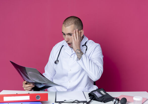 医生惊讶的年轻男医生穿着医用长袍和听诊器坐在办公桌旁 手里拿着工作工具 看着x光照片 手放在头上 隔离在粉红色的墙上拍摄穿着感觉