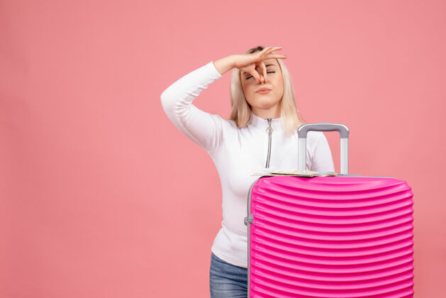 成人正面图：站在粉红色手提箱后面的年轻女士抱着鼻子站立女士前排