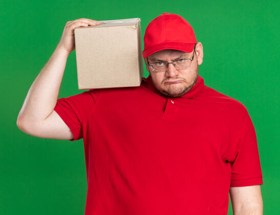 年轻恼怒的超重的年轻送货员戴着眼镜 肩上扛着纸板箱 隔离在绿墙上 留着复印空间光学手持恼火
