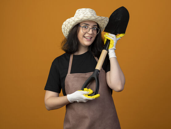 姿势惊讶的是 年轻的深色皮肤的女园丁戴着眼镜 穿着制服 戴着园艺帽和手套 把铲子孤立地放在橙色的墙上花园女性女人