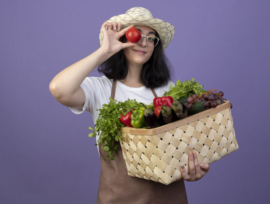 拿着年轻的深色皮肤的女园丁戴着眼镜 穿着制服 戴着园艺帽 手里拿着菜篮子 眼睛上涂着隔离在紫色墙上的西红柿脸姿势人