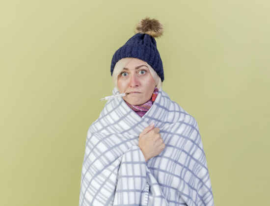 包裹震惊的年轻金发病女人戴着冬天的帽子和围巾 嘴里拿着用格子布包裹的温度计 隔离在橄榄绿的墙上嘴举行疾病