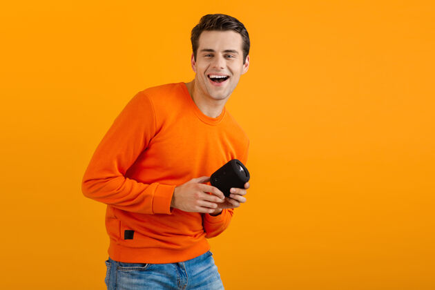 娱乐穿着橙色毛衣的时髦微笑的年轻人拿着无线扬声器快乐地听音乐享受橙色的乐趣肖像时尚男人