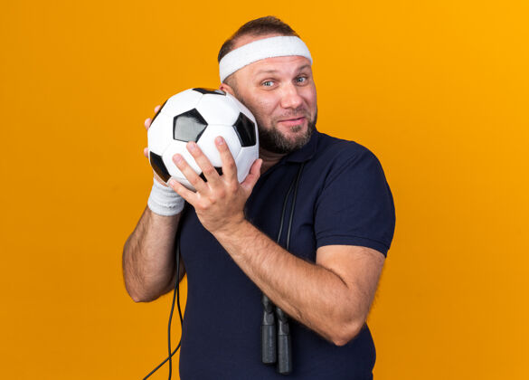 绳子高兴的成年斯拉夫运动男子与跳绳脖子上戴着头带和腕带举行的橙色墙上的复制空间隔离球运动男人抱着