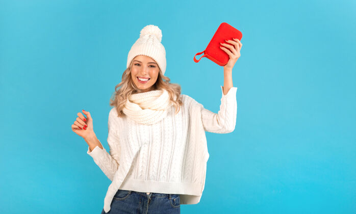 女性时尚的金发微笑的美丽年轻女子手持无线扬声器听音乐穿着白色毛衣和针织帽子摆在蓝色休闲温暖圣诞节