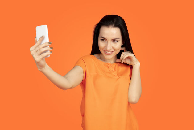工作橙色工作室背景上的白种人年轻女子肖像穿着衬衫的漂亮深色女性模特人类情感的概念 面部表情 销售 广告广告空间自拍 赌赢年轻人类衬衫