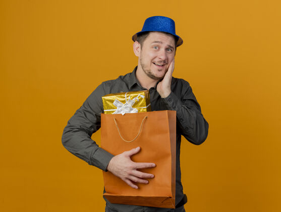 抱高兴的年轻人戴着蓝帽子 拿着礼品袋 把手放在脸颊上孤立的橙色礼物脸颊橙色