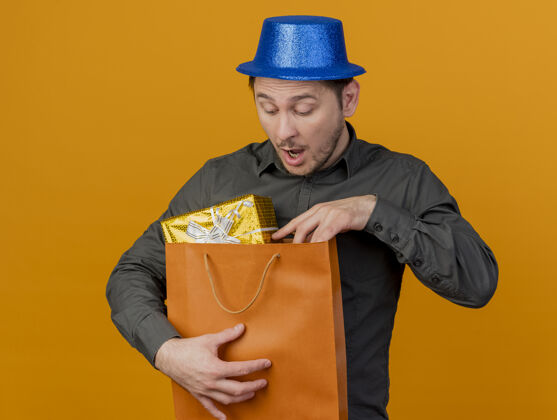 举行惊讶的年轻人戴着蓝色的帽子 拿着礼物袋看着孤立的橙色穿男人年轻人