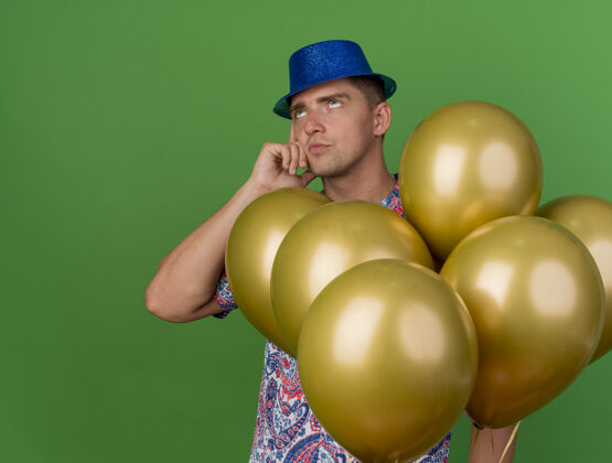 帽子一个戴着蓝帽子 体贴的年轻人站在绿色的气球中间在其中穿派对
