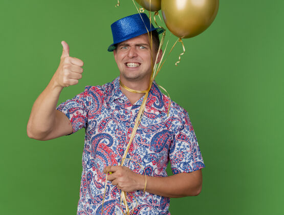 抱着愤怒的年轻人戴着蓝帽子 脖子上绑着气球 在绿色的草地上竖起大拇指帽子脖子围着