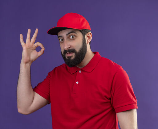 请高兴的年轻帅气的送货员穿着红色制服 戴着帽子 在紫色的墙上做着“ok”的牌子人Ok人