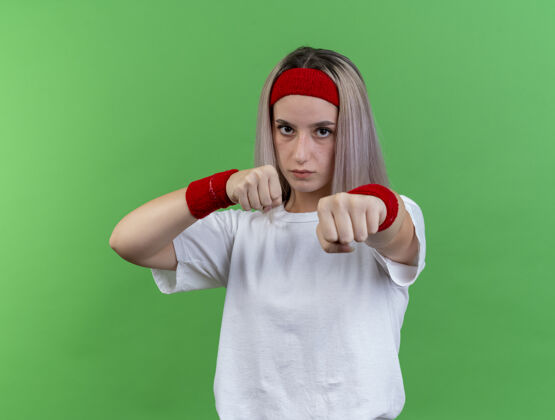 信心自信的年轻运动女性戴着背带 戴着头带和腕带 拳头随时准备在绿色的墙壁上击打拳打头
