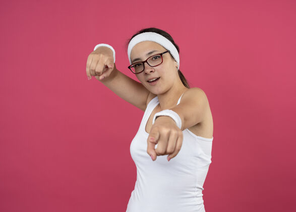 光学令人印象深刻的年轻运动女性戴着眼镜戴着头带和腕带指着前面的粉红色墙壁上孤立女印象人
