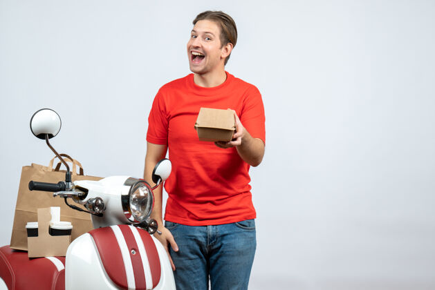 抱身着红色制服 面带微笑的送货员站在踏板车旁 手里拿着白色背景的小盒子运动滑板车小