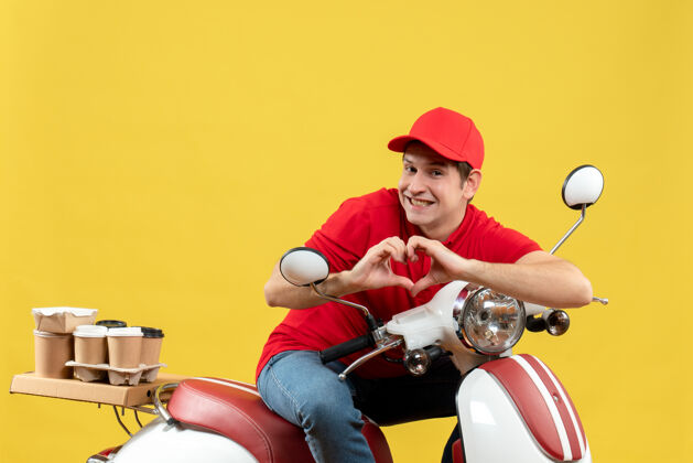 红顶视图浪漫的年轻人穿着红色衬衫和帽子传递命令 使黄色背景上的心脏姿态帽子摩托车年轻人