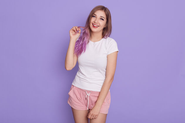 笑话一个穿着白色t恤和玫瑰色短发的快乐欢笑的年轻女孩 表情调情 手指放在淡紫色的头发上 在紫色的墙上摆出孤立的姿势乐观牙牙学语积极